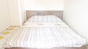 Ein Einzelbett mit gestreifter Bettdecke und einem Holz-Kopfstück in einem Zimmer mit weißer Tür und einem gelb-weißen Fliesenboden.