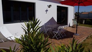 Una casa moderna con un patio que tiene una mesa y sillas, un paraguas rojo y un pequeño jardín con plantas.