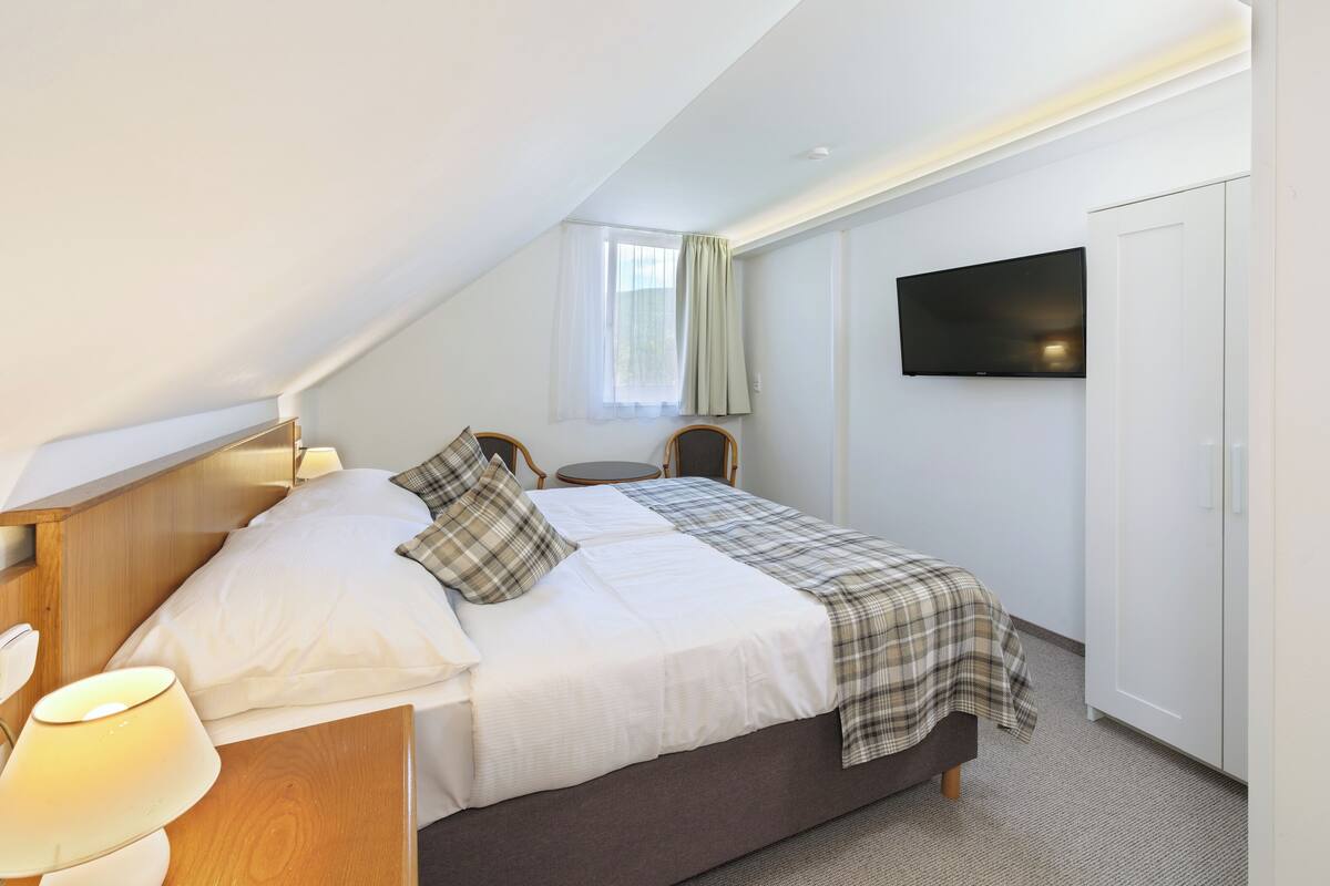 A hotel room with a bed, a bedside table with a lamp, a TV mounted on the wall, and a window with curtains.