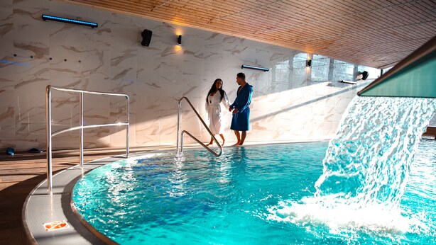A swimming pool with a marble wall backdrop, a metal ladder, and a water feature. - Wellness Hotel Astra (Spindleruv Mlyn)
