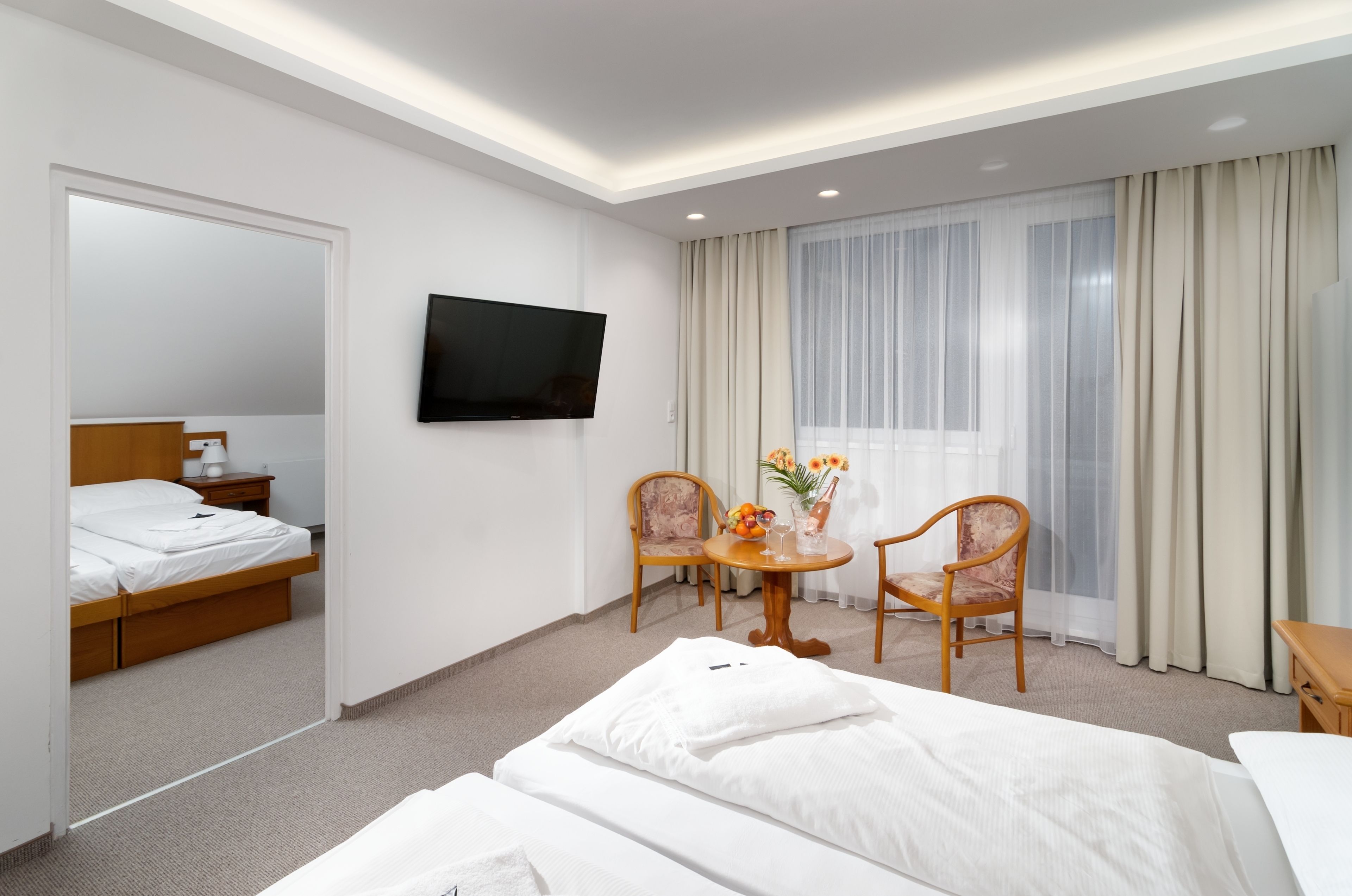 A hotel room with a bed, a TV mounted on the wall, a small table with chairs, and a window with curtains.