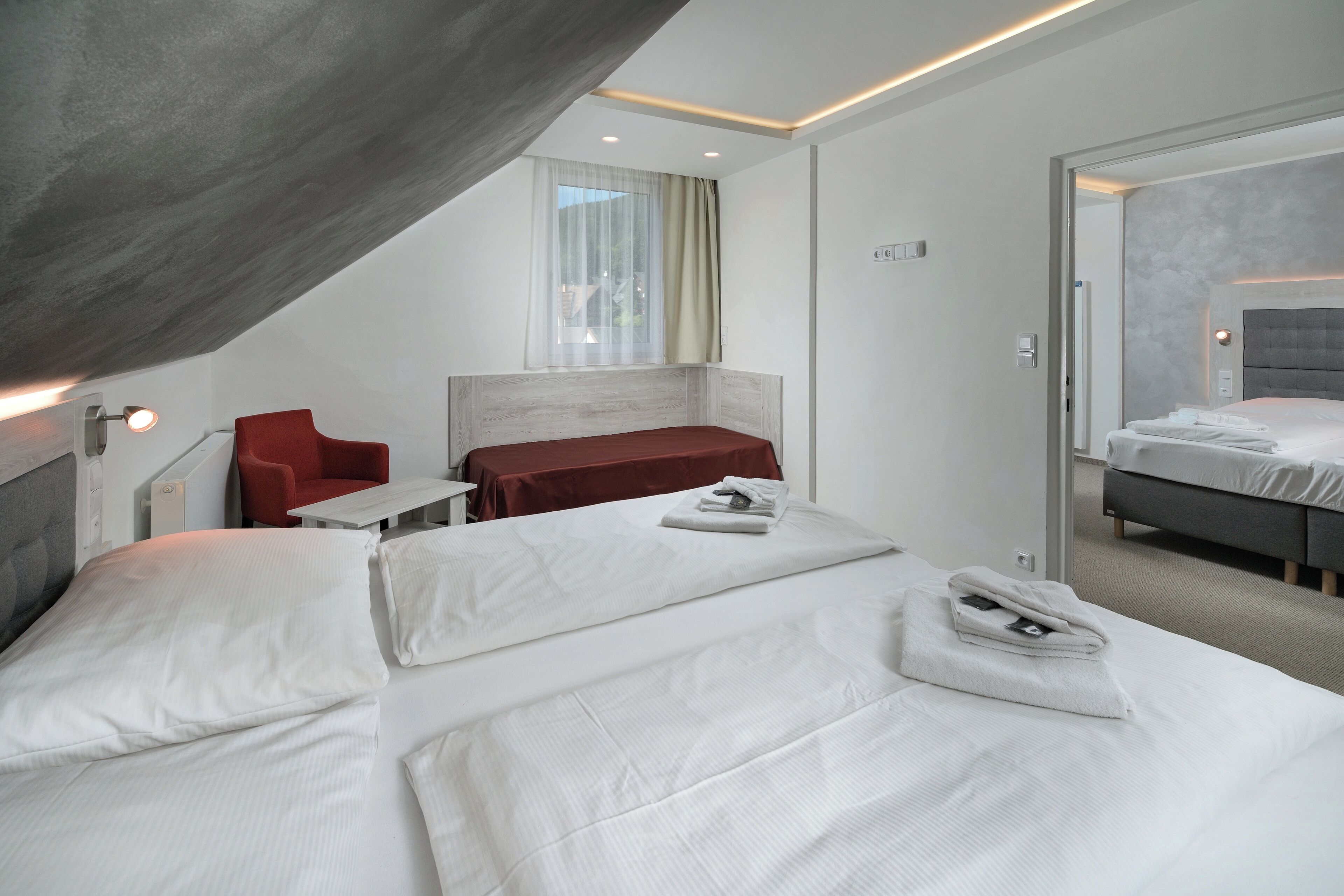 A modern hotel room with two beds, a red chair, a small table, and a large window with curtains.
