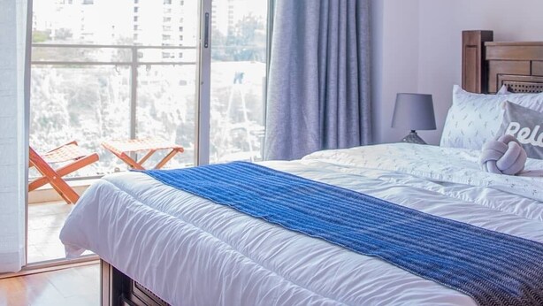 A hotel room with a large window, a bed with a blue striped blanket, and a view of buildings outside. - Spacious one bedroom apartment in Kileleshwa Nairobi (Nairobi)