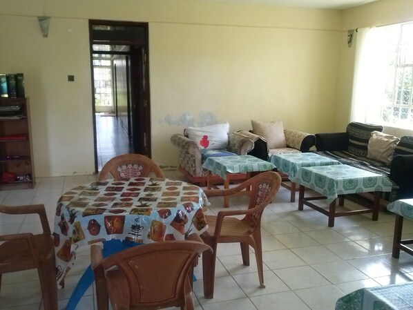 A dining area with a table covered in a patterned tablecloth, surrounded by wooden chairs. There is a sofa set with patterned cushions and a window with a view.