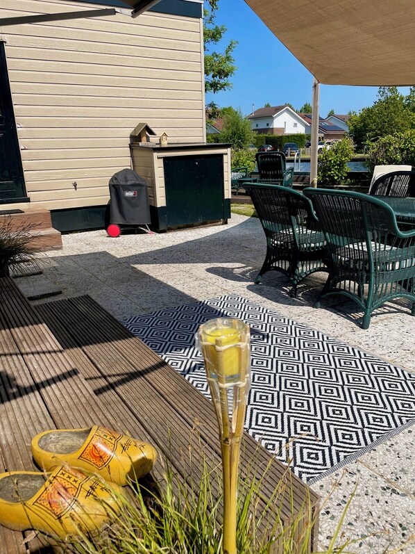 Un espace extérieur aménagé en terrasse, avec un sol à motifs, des chaises vertes, un barbecue et une table couverte.