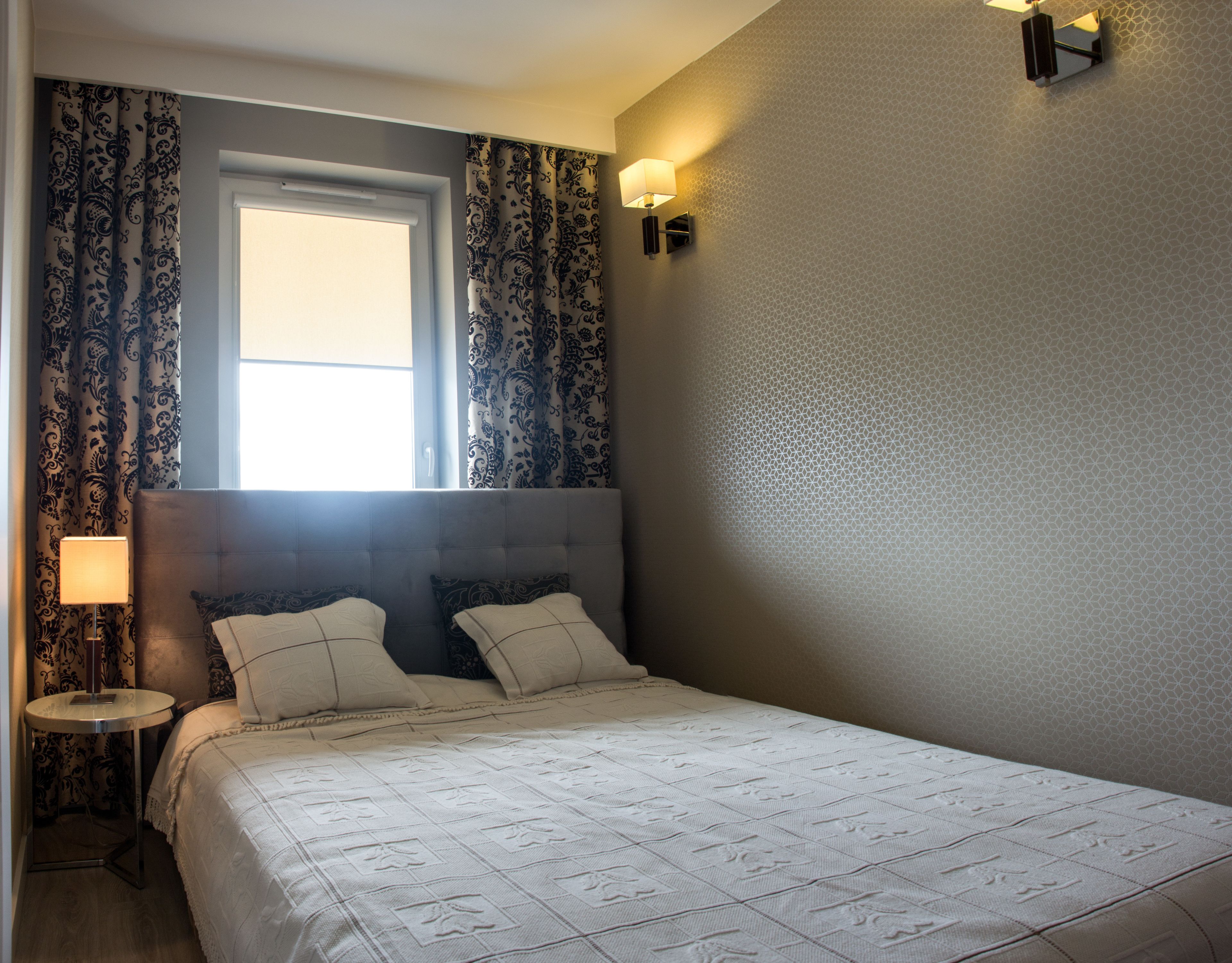 A bedroom with a large bed, two bedside lamps, a window with curtains, and a wall-mounted light fixture.