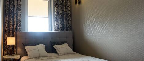 A bedroom with a large bed, two bedside lamps, a window with curtains, and a wall-mounted light fixture.