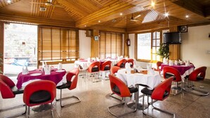 A conference room with wooden ceiling and walls, multiple tables with red chairs, and large windows. - Hotel Aangan Regency (Shimla)