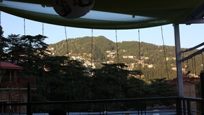 A view from a balcony with a hanging decoration, a railing, and a distant village on a hillside. - Hotel Aangan Regency (Shimla)