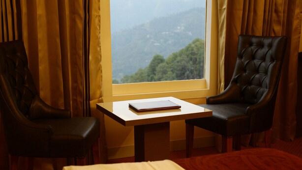 A room with a bed, two leather chairs, a small table with a notebook, and a window with a view of a forested mountain. - Hotel Aangan Regency (Shimla)