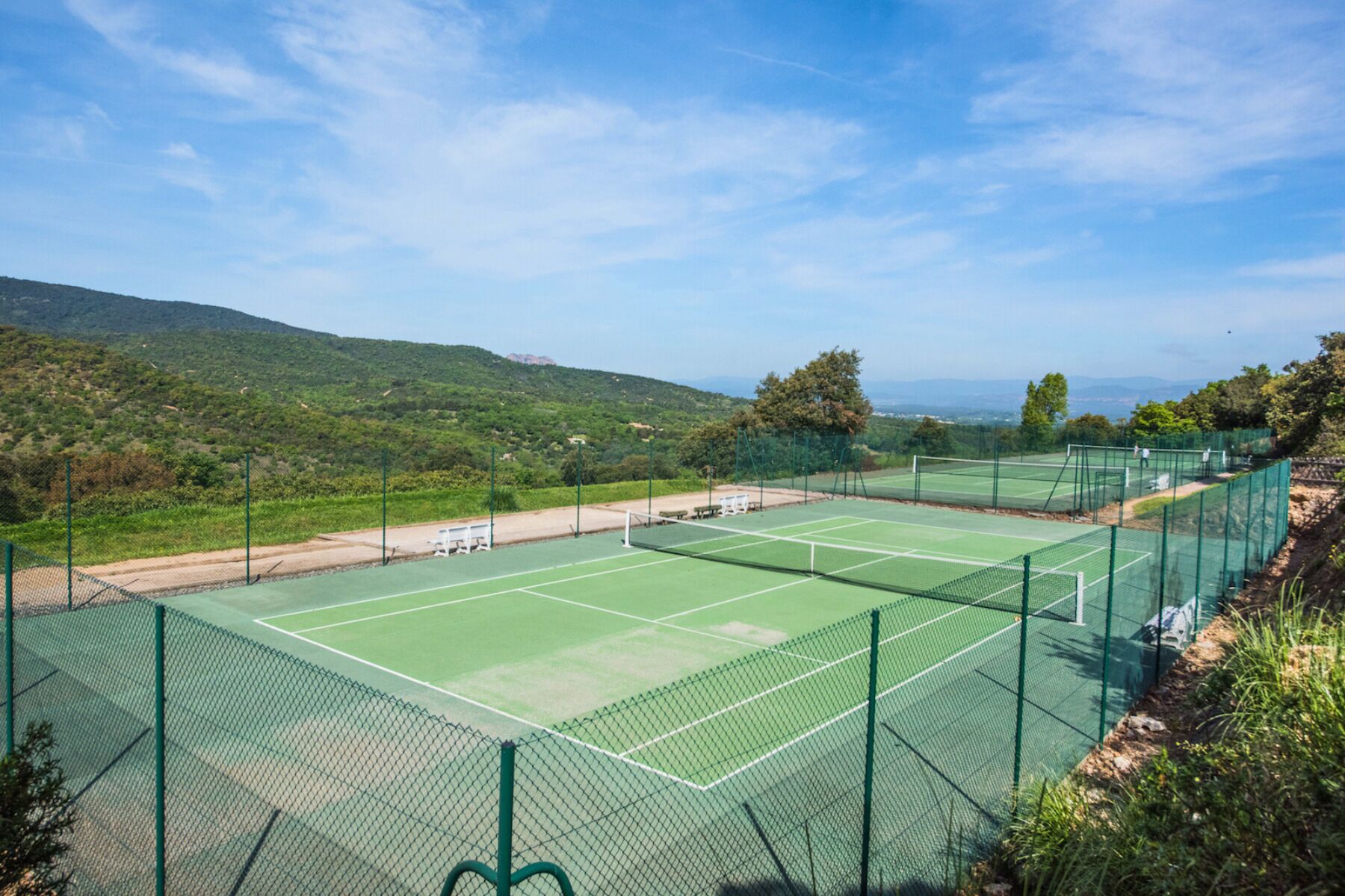 Ein Tennisplatz mit Netz, umgeben von einem grünen Zaun, mit Hügeln und blauem Himmel im Hintergrund.