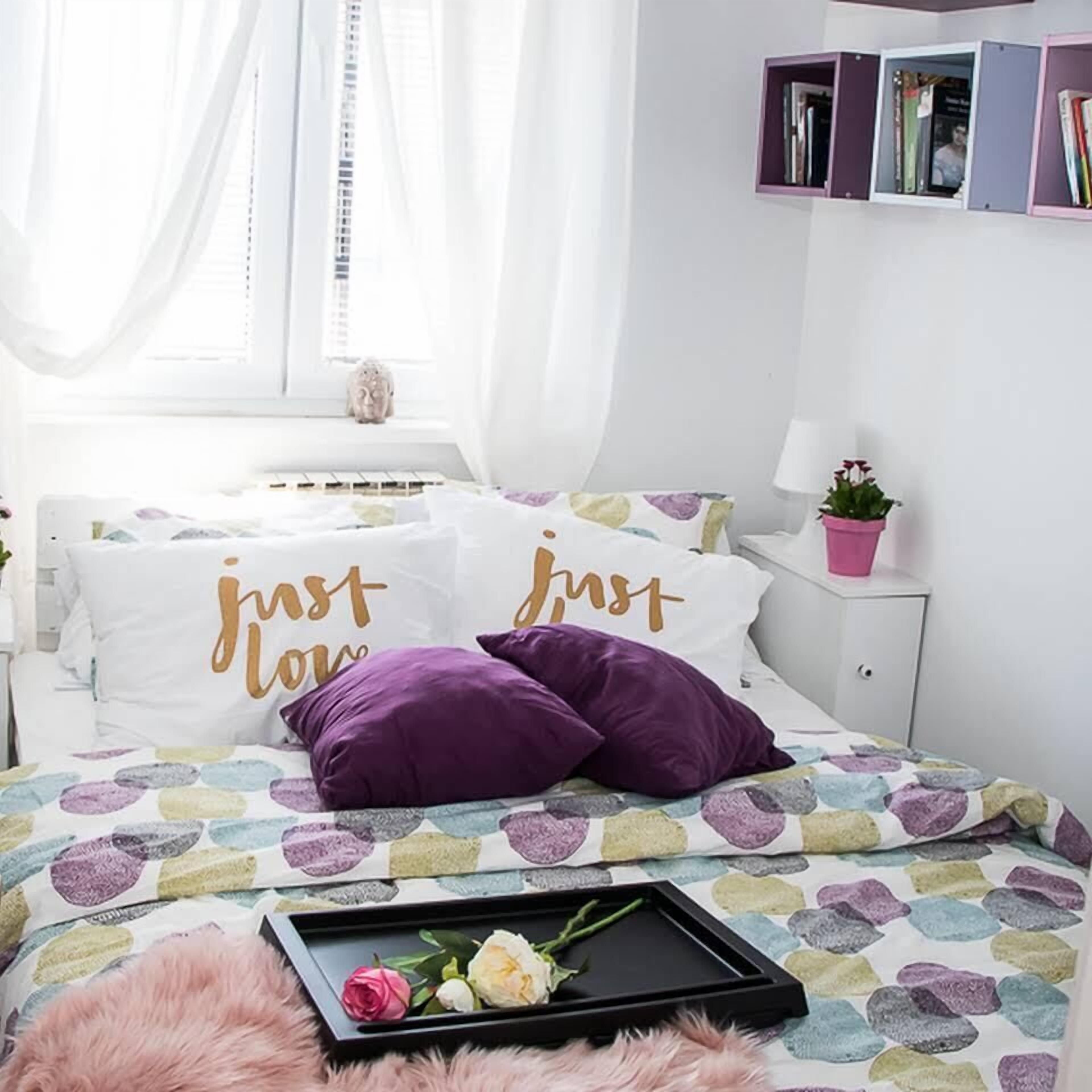 A bedroom with a bed, pillows, a bedside table with a lamp, a window with curtains, a small statue, and a bookshelf.