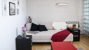 Una sala de estar moderna con un sofá blanco, cojines blanco y negro, una manta roja, una mesita de noche con una planta y una lámpara colgante.