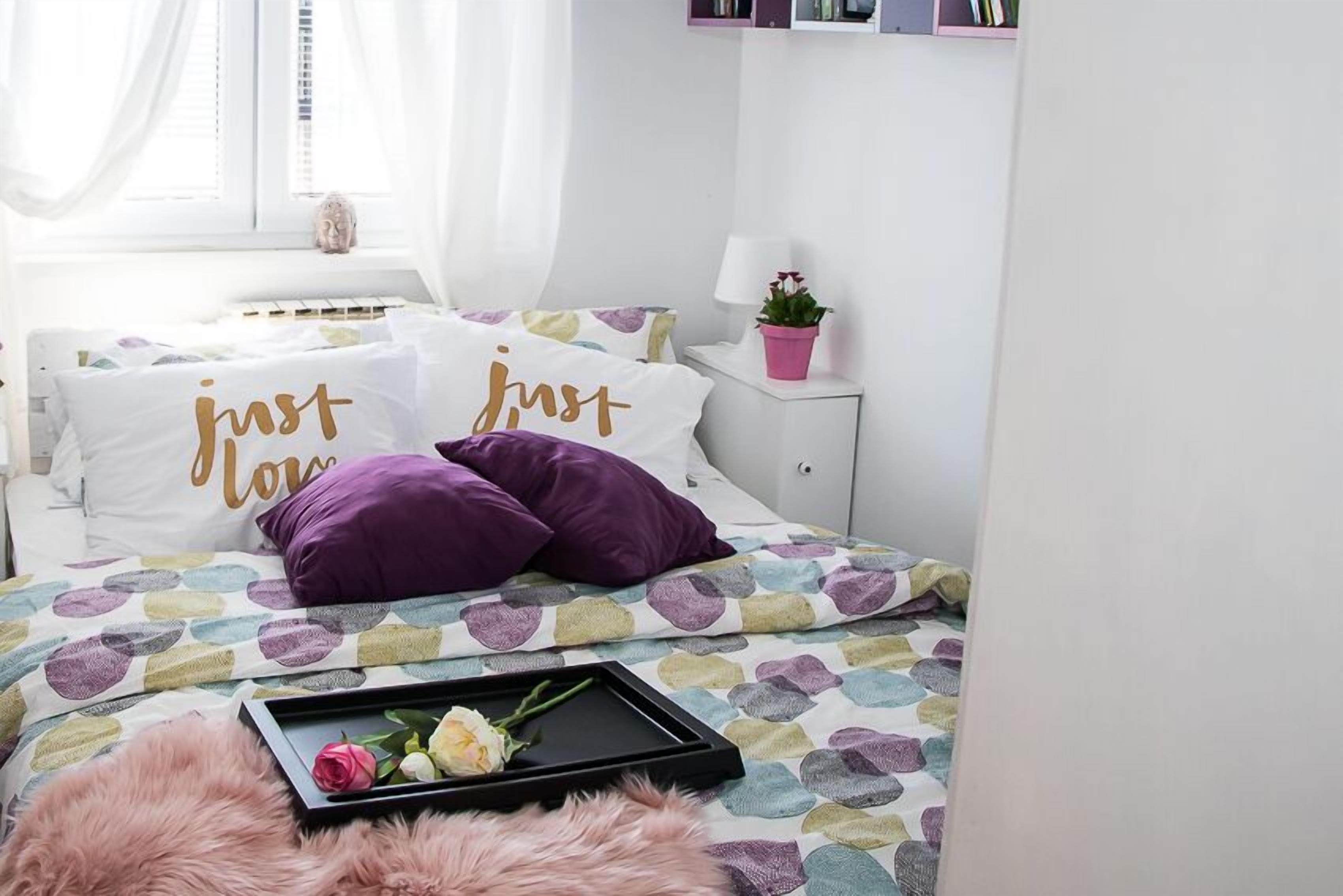 Una cama bien hecha con ropa de cama estampada, una bandeja con una rosa y una ventana con cortinas traslúcidas.