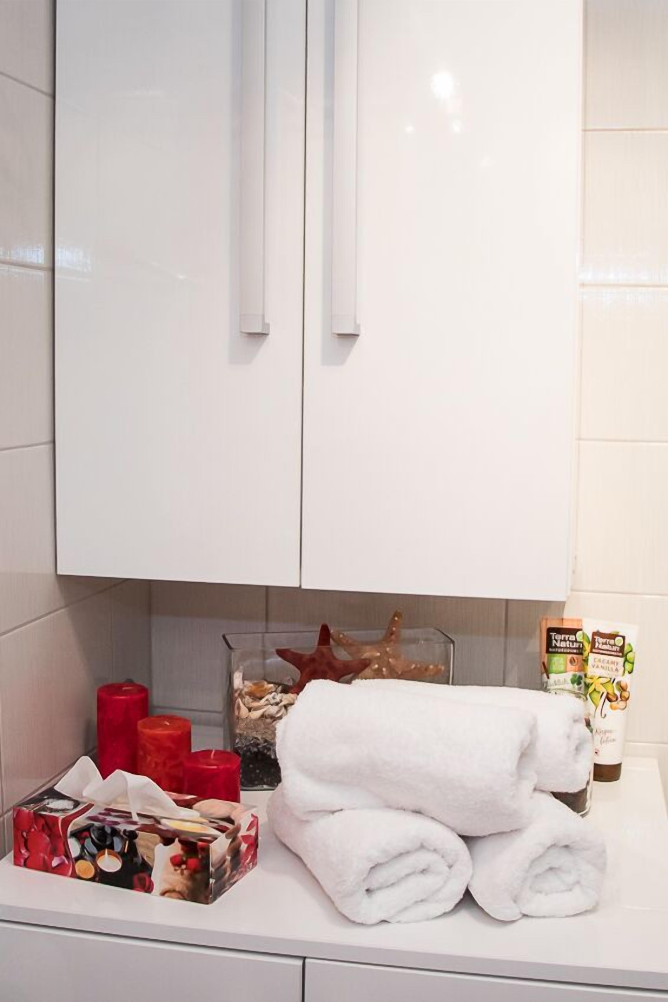 Une salle de bain avec des meubles blancs, un bocal en verre, des bougies rouges, une boîte de mouchoirs et une serviette blanche roulée posée sur le comptoir.