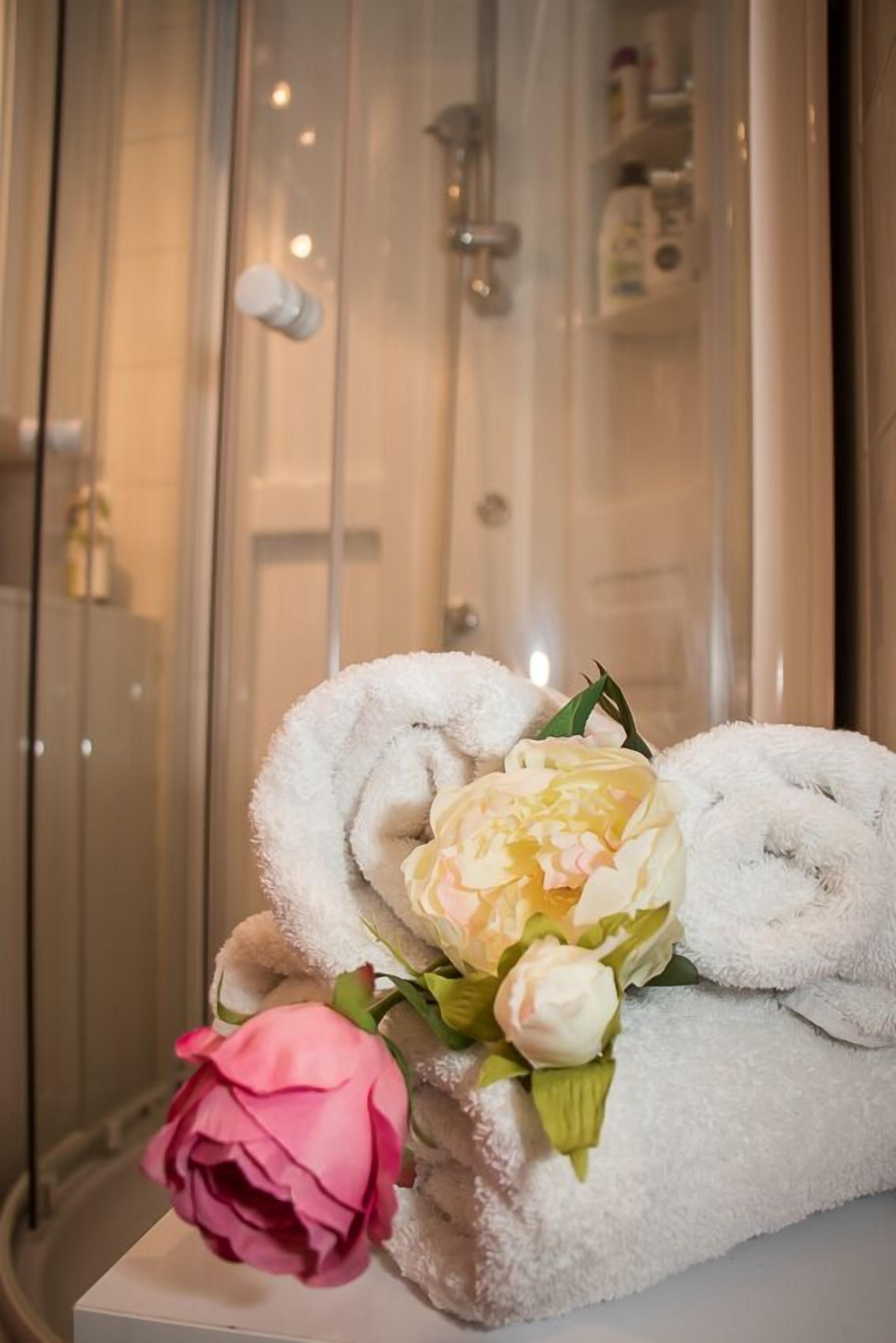 A bathroom with a glass shower enclosure, a towel rack with folded towels, and a decorative arrangement of flowers.