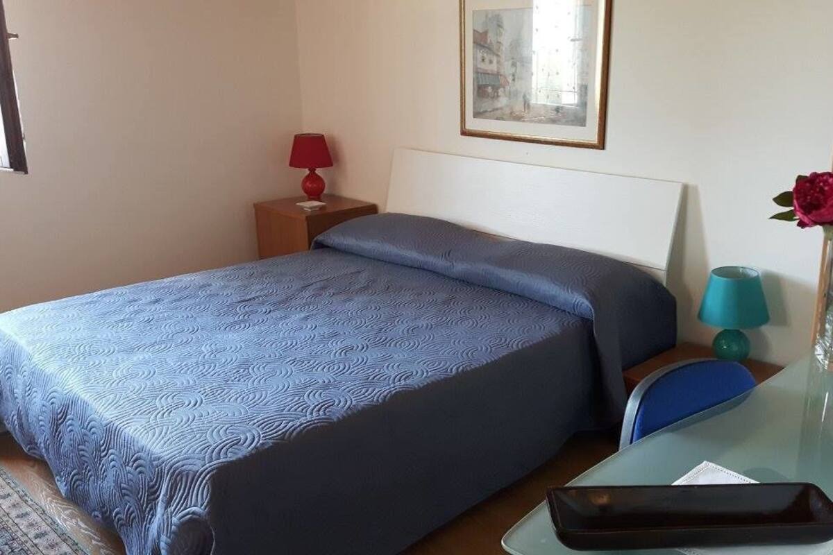 A hotel room with a bed, a nightstand with a lamp, a table with a vase of flowers, and a chair.