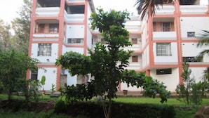 A multi-story building with a white facade and pink accents, surrounded by greenery and a small tree in the foreground. - Snow Peak Hotel (Chogoria)