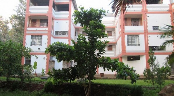 A multi-story building with a white facade and pink accents, surrounded by greenery and a small tree in the foreground.