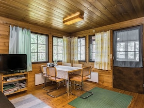 A wooden cabin interior with a dining table, chairs, a television, and a bookshelf.