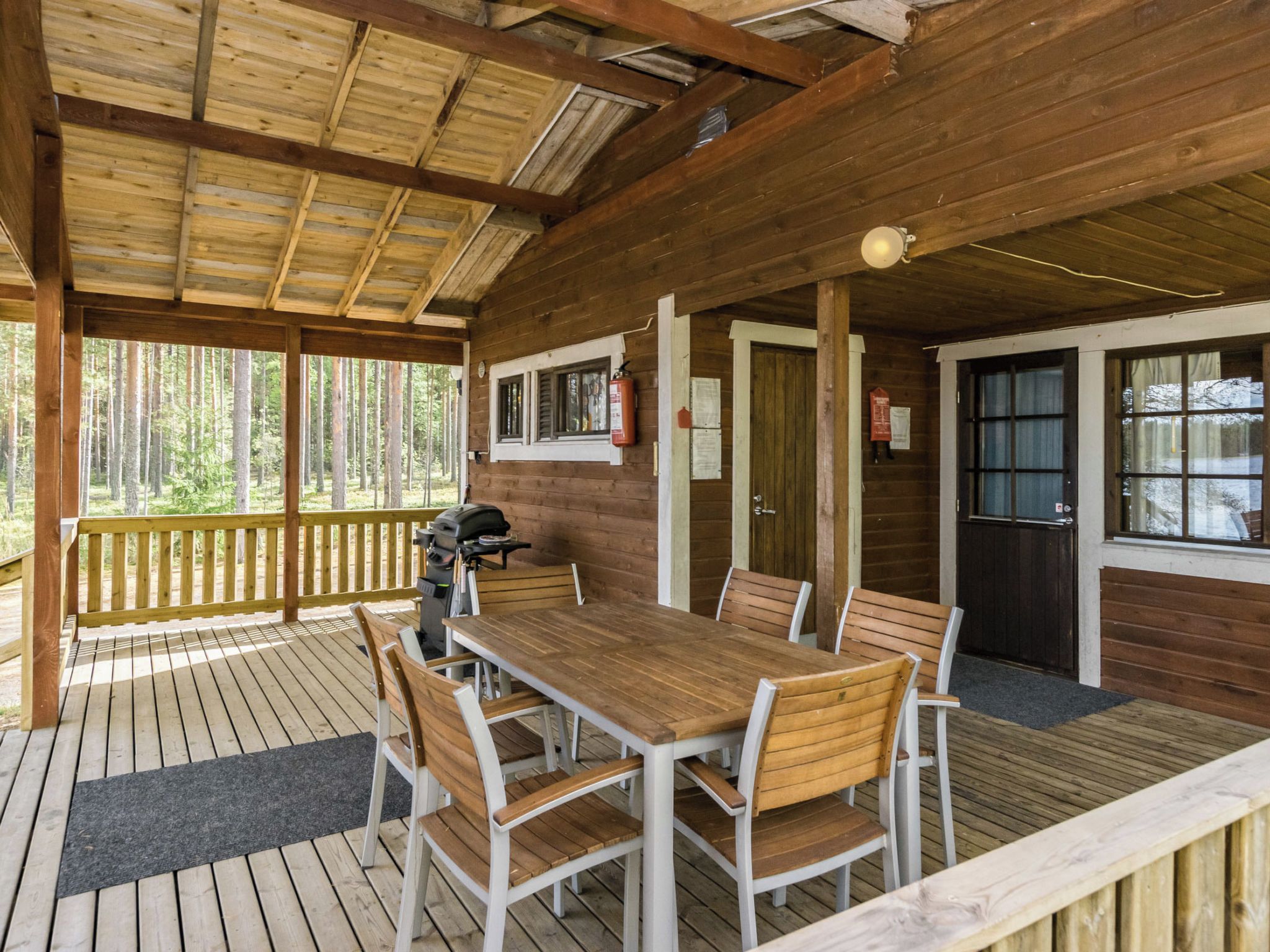 A covered outdoor dining area with a wooden table and chairs, a grill, and a view of the forest.