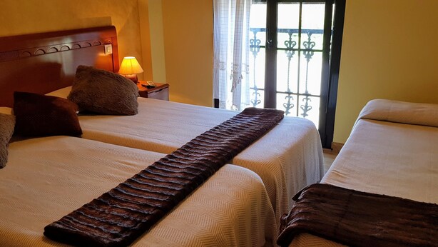 Une chambre d’hôtel avec deux lits, une table de chevet avec une lampe et une vue par une fenêtre ornée de barreaux en fer décoratifs.