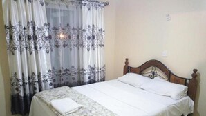 A neatly made bed with a floral bedspread and white linens, a wooden headboard, and sheer curtains with a floral pattern. - Muthama Heights (Syokimau)