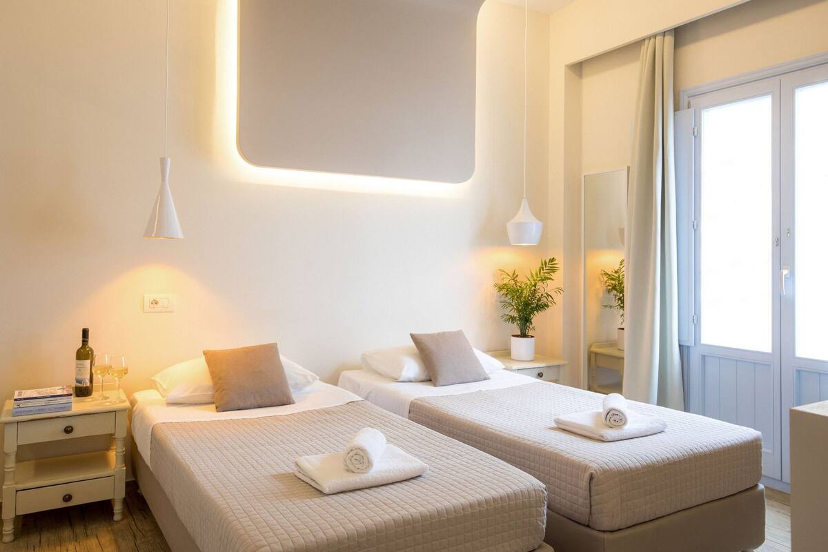 A hotel room with two beds, a nightstand with a bottle and glasses, and a potted plant.