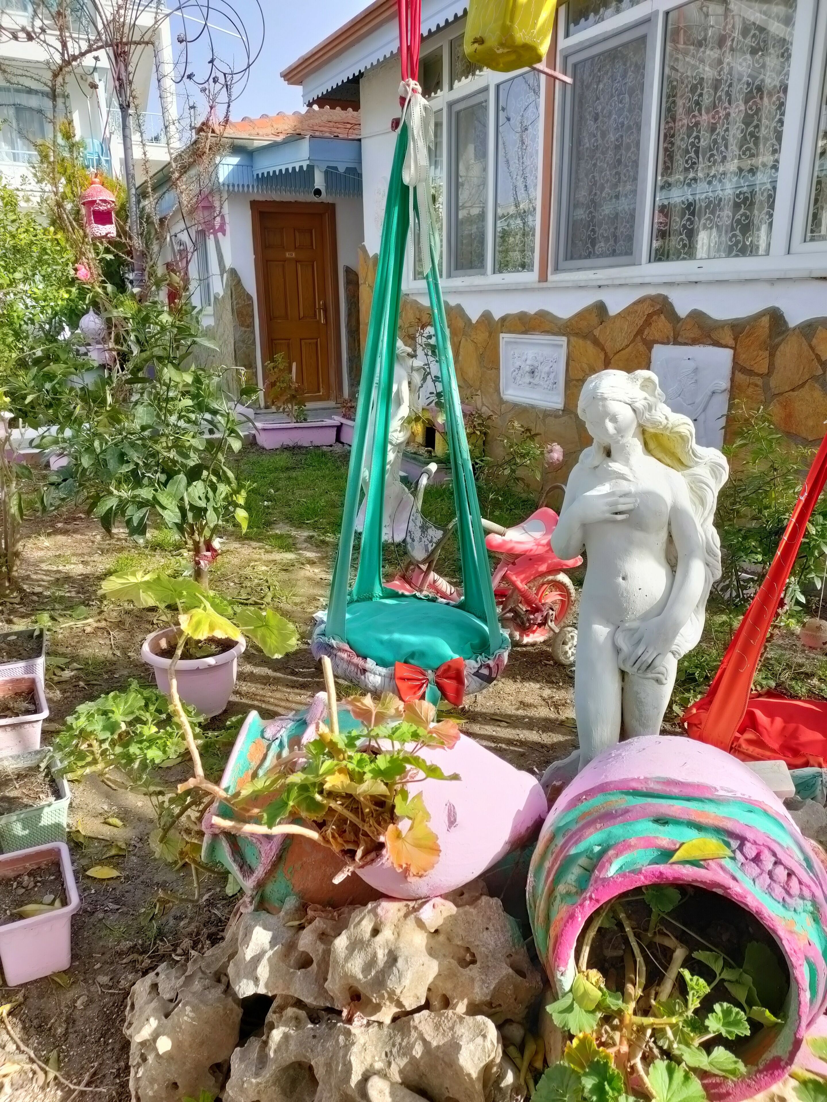 Un jardín con una hamaca colgante, una estatua y diversas macetas con plantas.