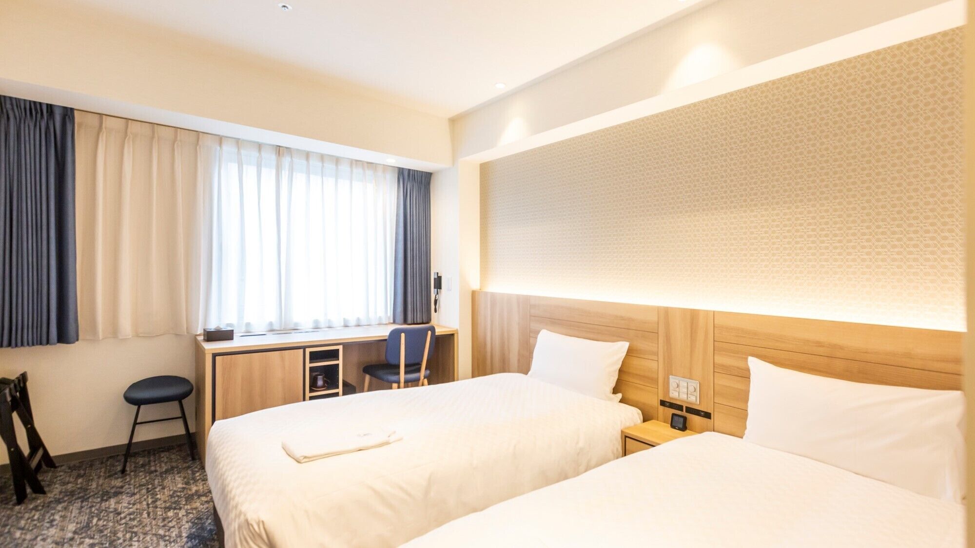A hotel room with two beds, a desk, a chair, and a large window with curtains.