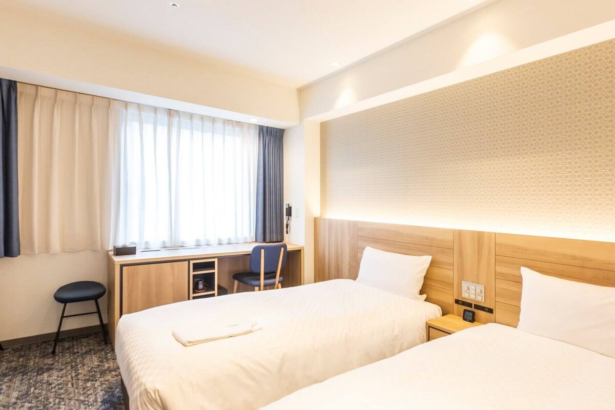 Habitación de hotel con dos camas, un escritorio, una silla y un ventanal con cortinas.
