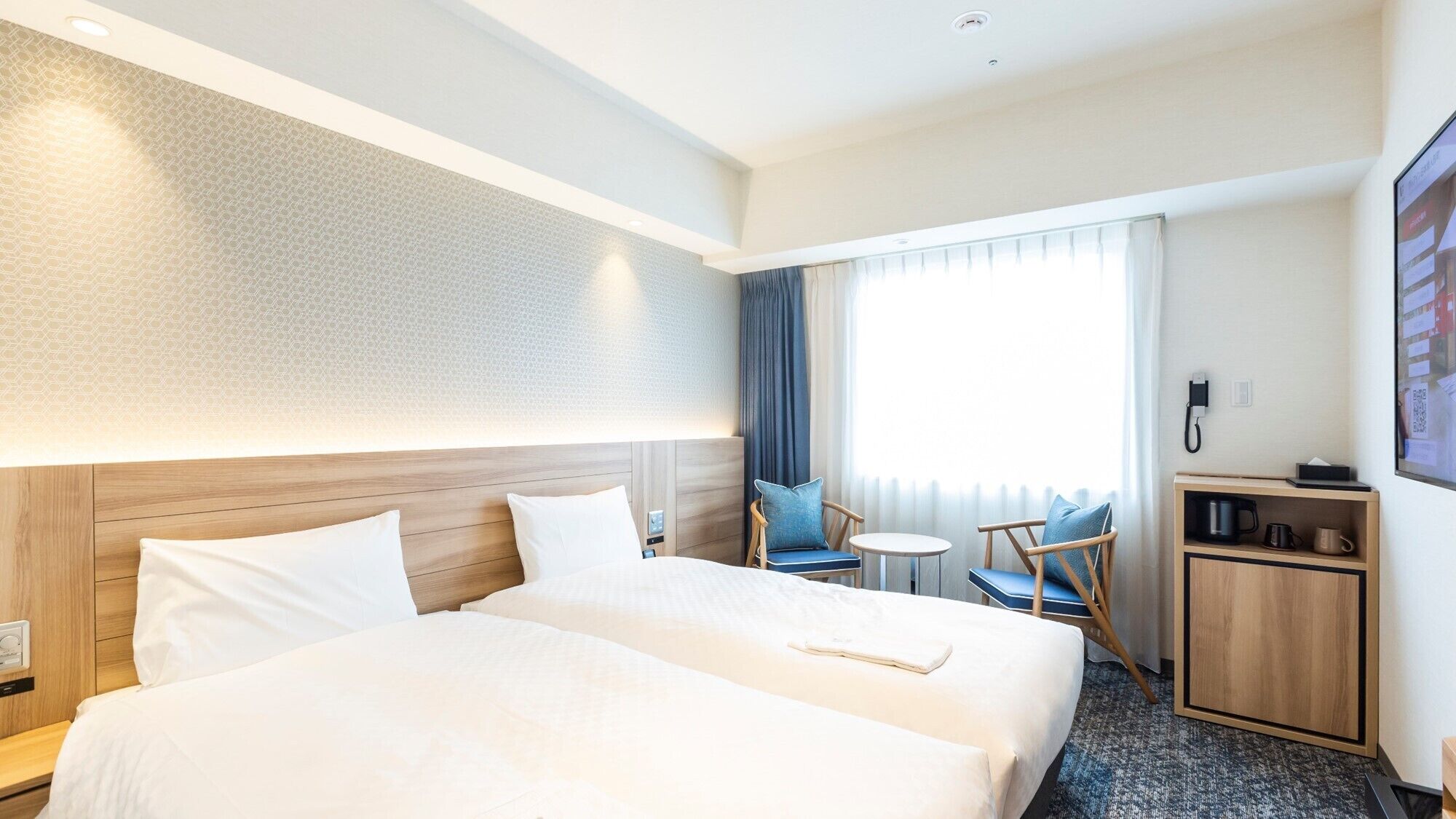 A hotel room with two beds, a small table, a chair, a desk, and a window with curtains.