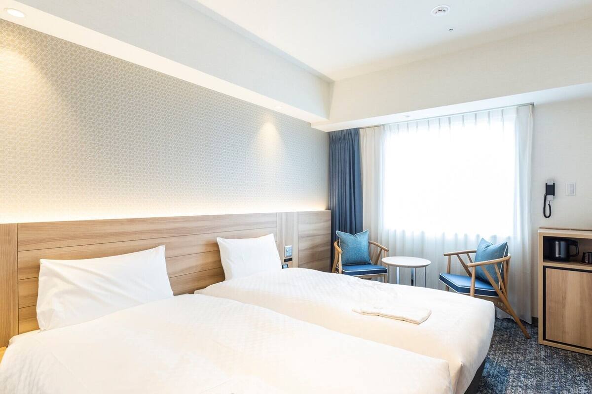 Habitación de hotel con dos camas, una mesita, una silla, un escritorio y una ventana con cortinas.