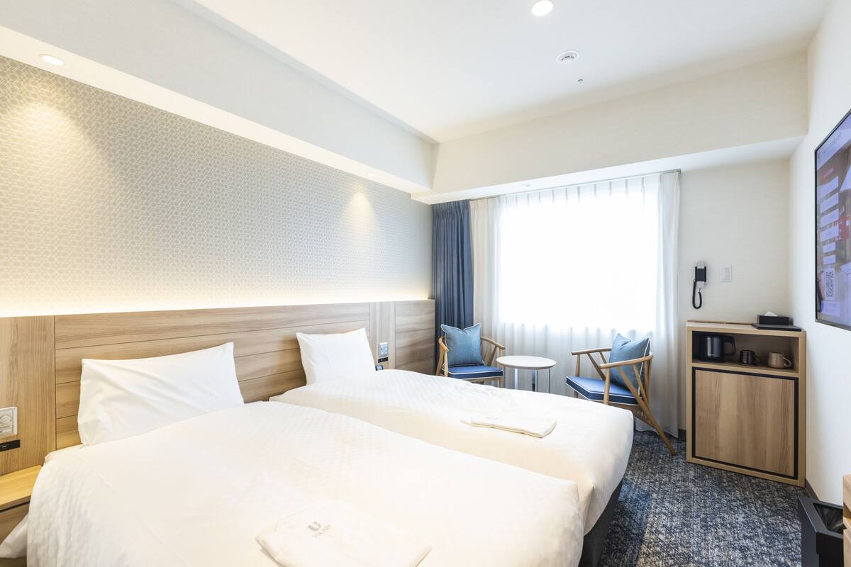 Habitación de hotel con dos camas, una mesita pequeña, una silla, un armario y una ventana con cortinas.