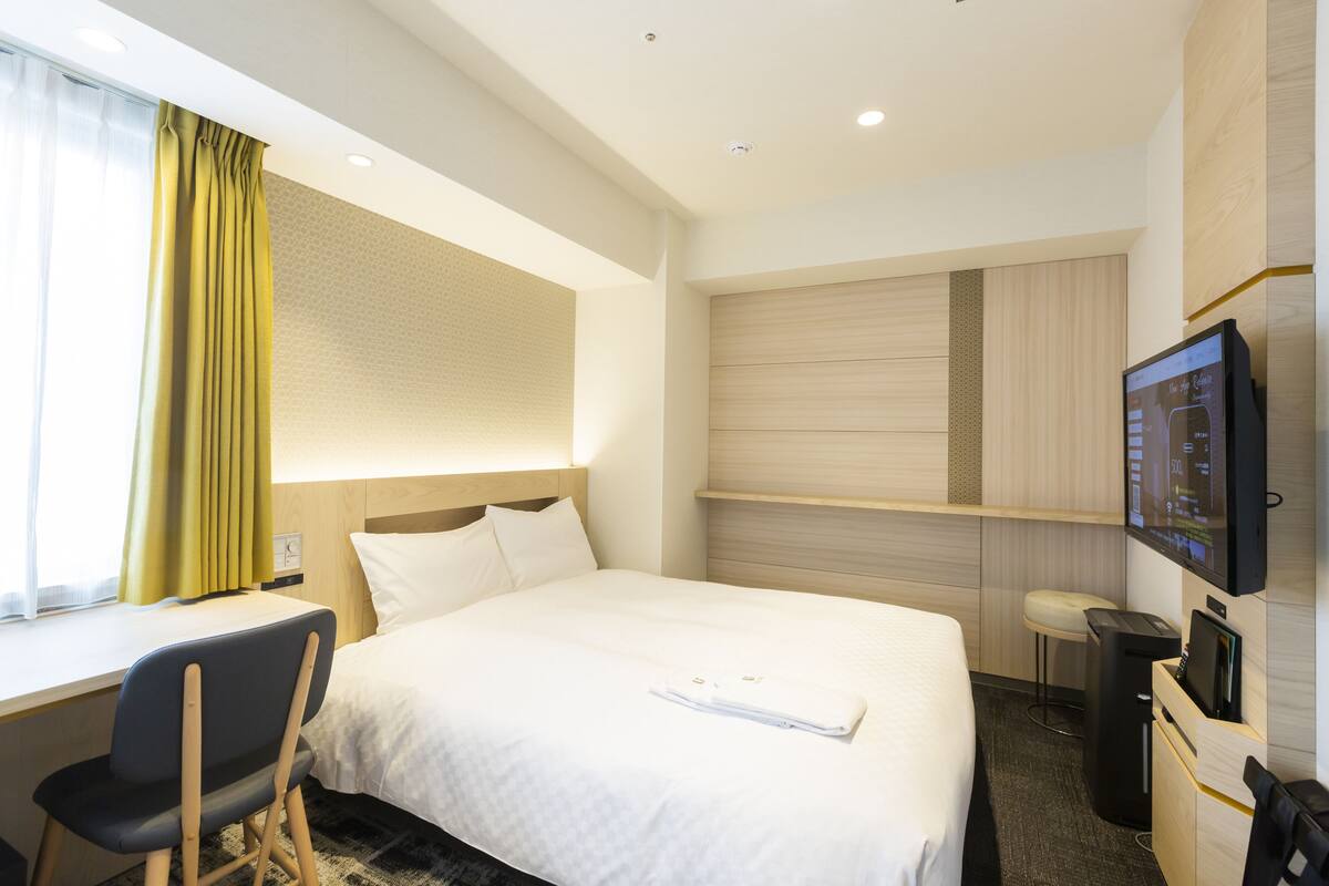 Una habitación de hotel con cama, escritorio, silla, televisor y ventana con cortinas.