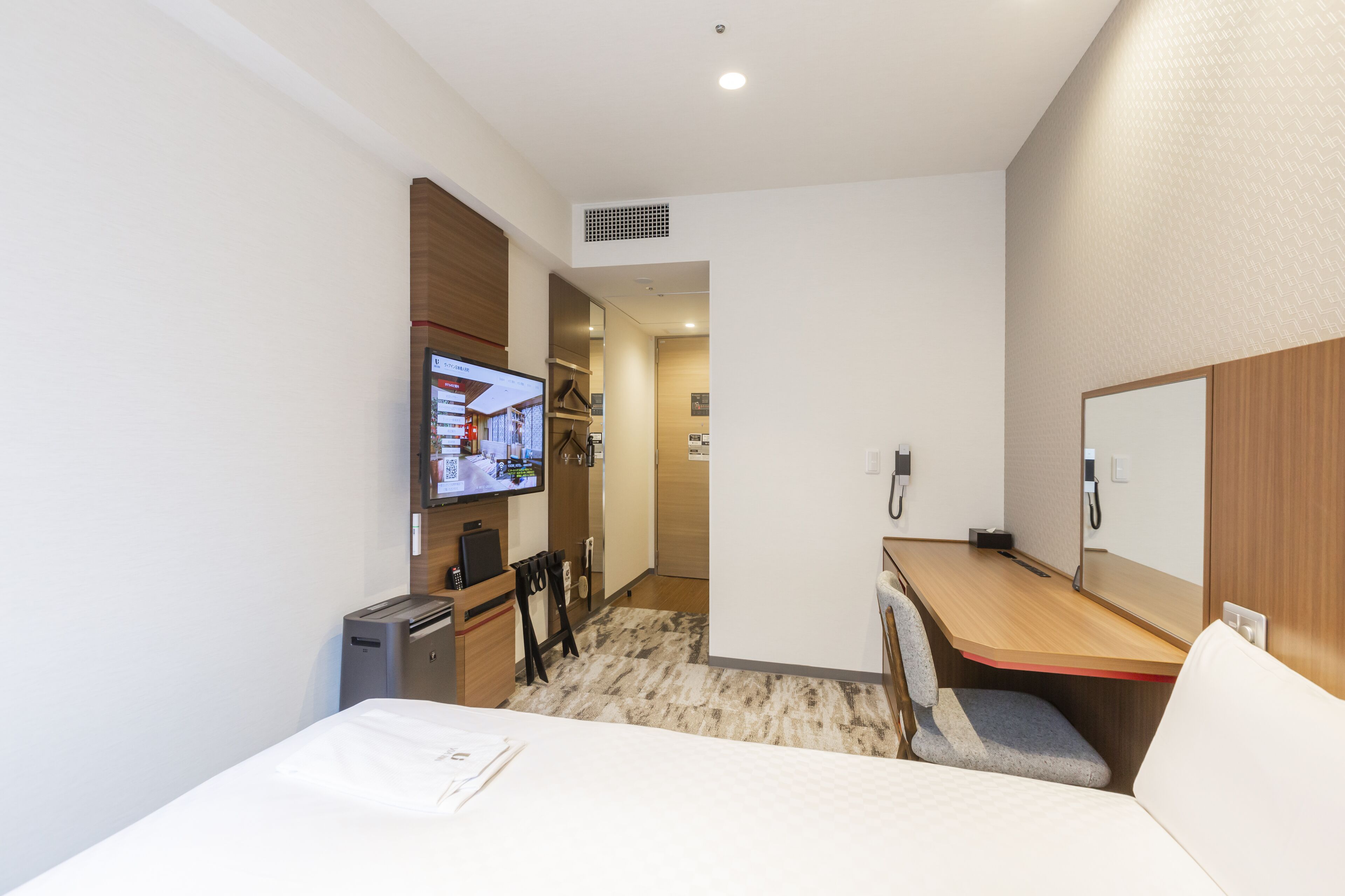 A compact hotel room with a bed, a desk, a chair, a television, and a small kitchenette.