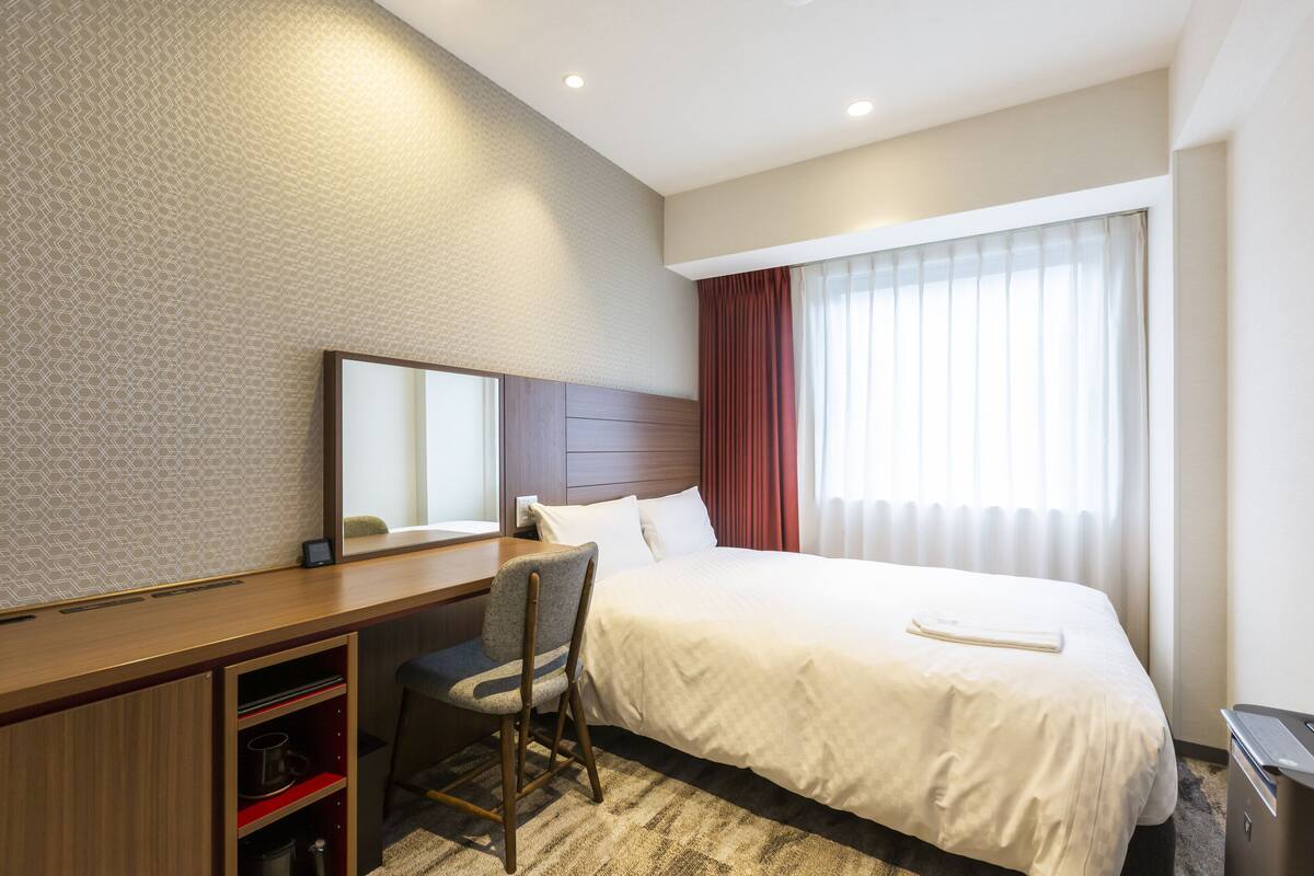 Habitación de hotel con cama, escritorio, silla, espejo y ventana con cortinas.