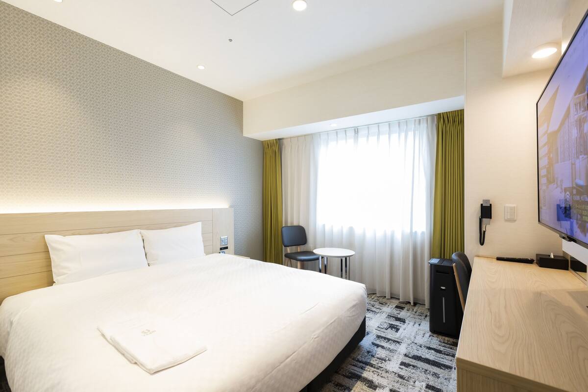 Una habitación de hotel con una cama grande, un escritorio, una silla, un televisor y una ventana con cortinas.