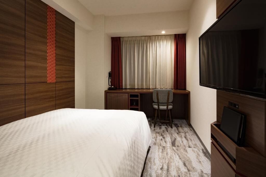 Quarto de hotel com uma cama, uma escrivaninha com cadeira, televisão e um guarda-roupa.