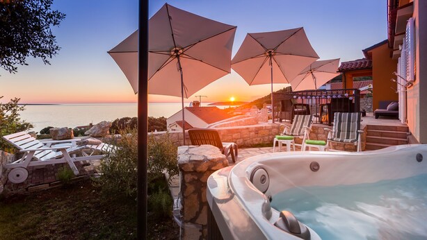 A hot tub with lounge chairs and umbrellas, overlooking a scenic ocean view at sunset.