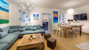 A modern living room with a sectional sofa, a wooden coffee table, and a dining area with a wooden table and chairs. There is a chandelier, artwork on the walls, and a TV mounted on the wall.