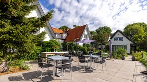 Een terras buiten met tafels en stoelen, omgeven door groen en gebouwen.