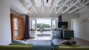 Een kamer met een groene bank, een houten kast, een televisie en uitzicht op een groen landschap door een groot raam.