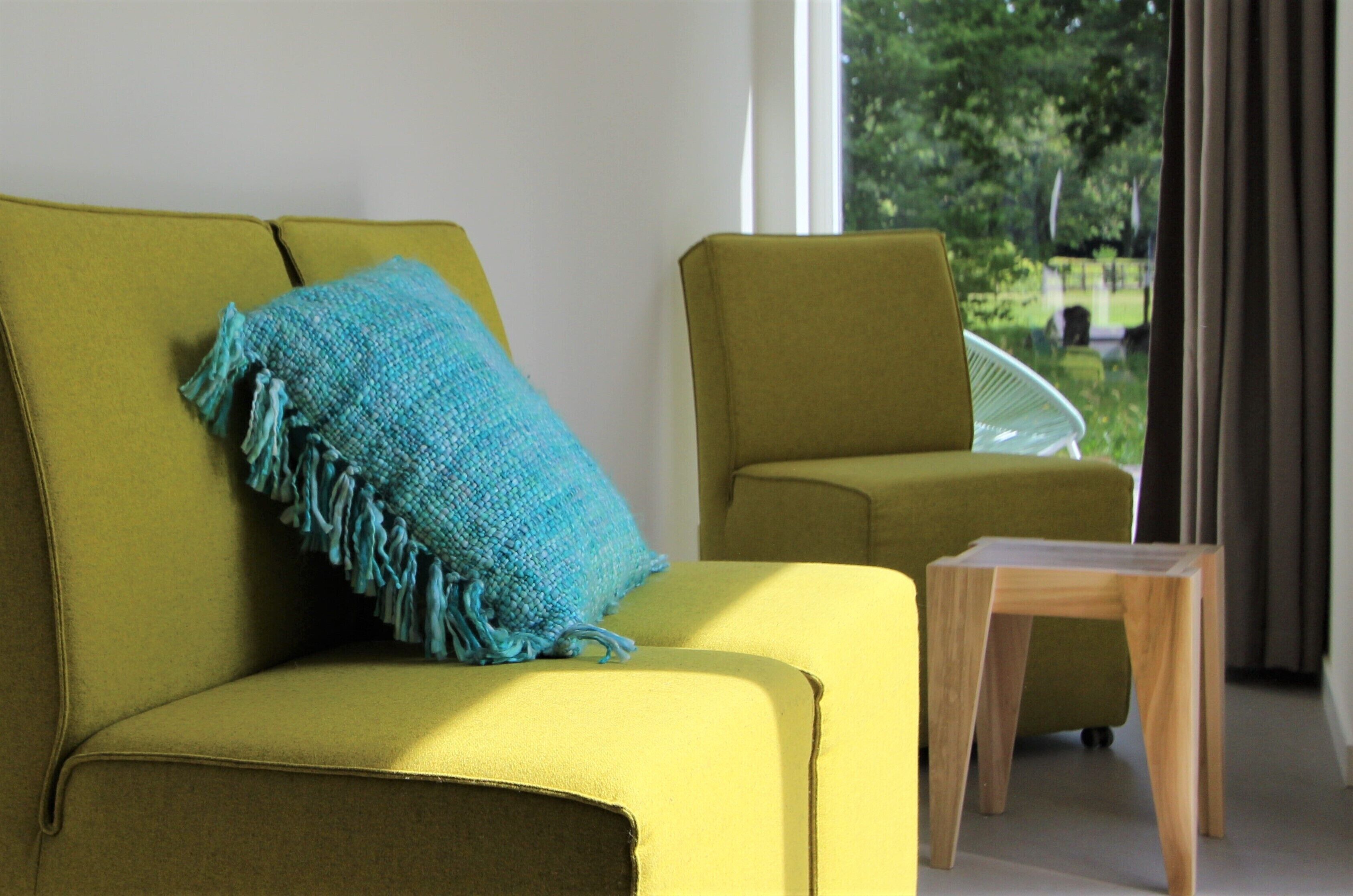 Un canapé jaune agrémenté d’un coussin bleu et d’une table d’appoint en bois, dans une pièce offrant une vue sur la verdure environnante.