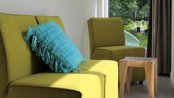 Un canapé jaune agrémenté d’un coussin bleu et d’une table d’appoint en bois, dans une pièce offrant une vue sur la verdure environnante.