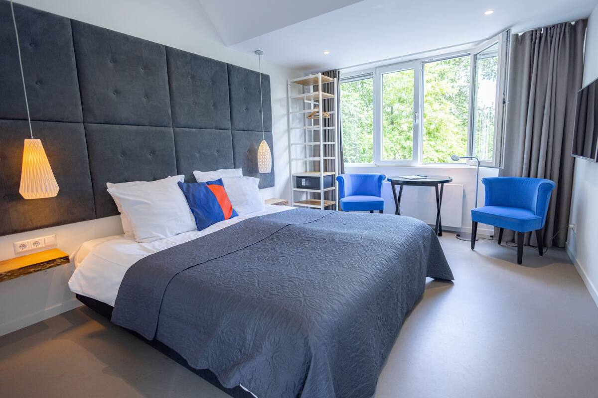 A modern hotel room with a large bed, a small table, two blue chairs, a TV, and a large window with a view of trees.