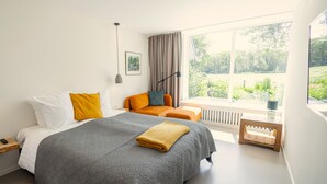 Een moderne slaapkamer met een groot bed, een kleine zithoek, een raam met uitzicht op groen en een nachtkastje met een lamp.