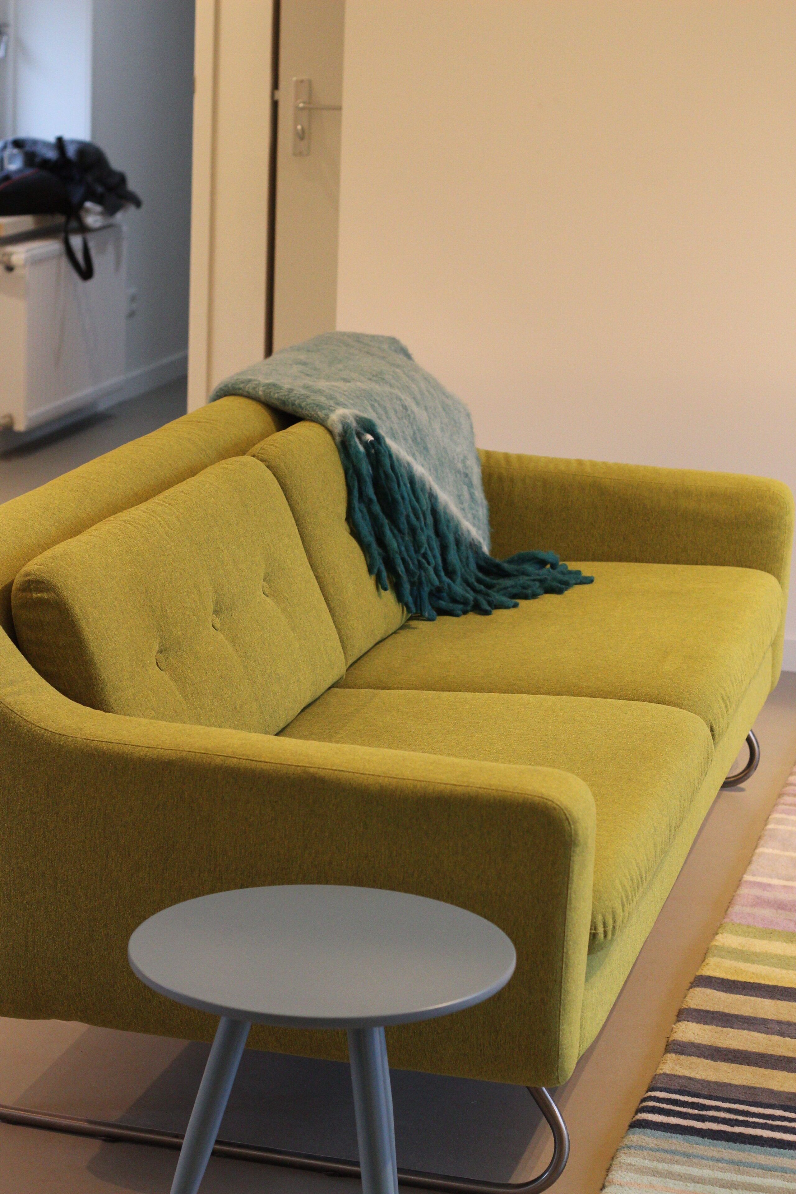 Un canapé jaune moutarde agrémenté d’un plaid turquoise, une petite table ronde et un tapis à rayures.