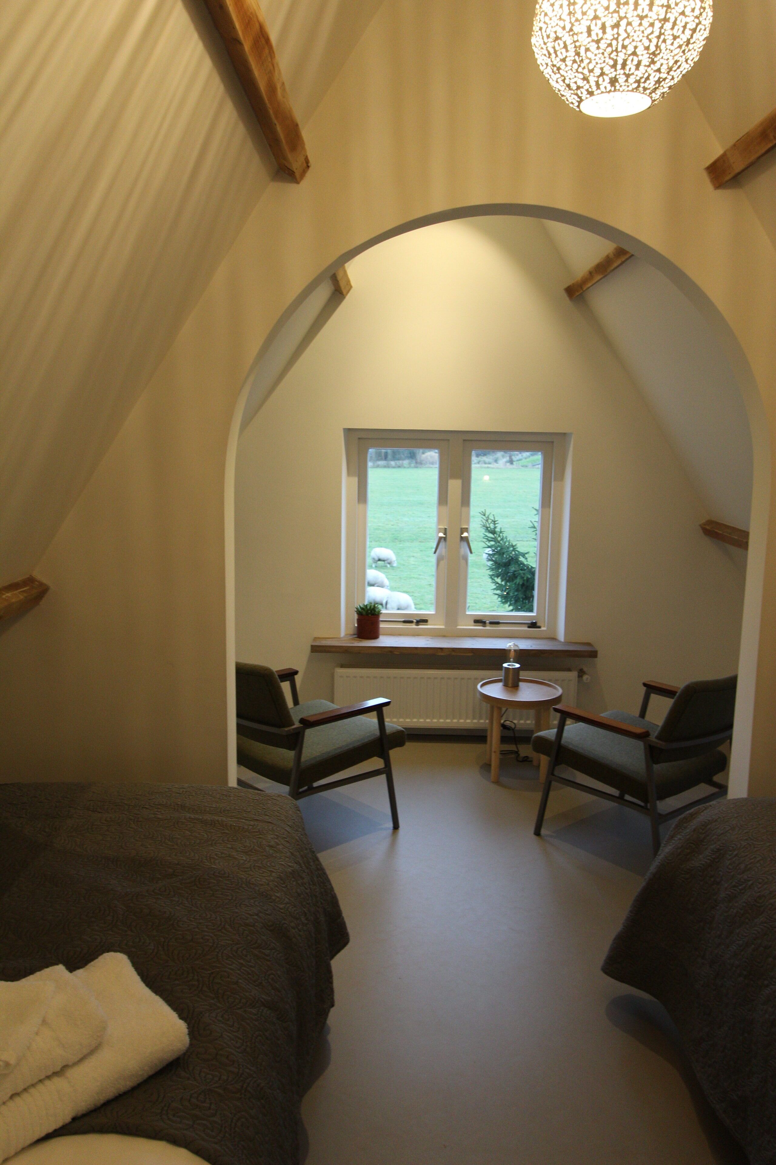A cozy attic bedroom with two beds, a small table, and a window overlooking a grassy field.