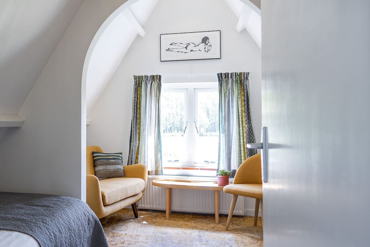 A bedroom with a bed, two chairs, a table, a hanging light fixture, a window with curtains, and a framed picture on the wall.
